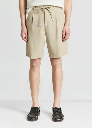 Mann trägt beige knielange Shorts mit Tunnelzug in der Taille und schwarze geschlossene Sandalen und steht vor einem weißen Hintergrund.
