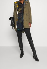 Parka verde olivo con forro de capucha esponjoso azul y amarillo, usada sobre una camisa oscura de flores, combinada con pantalones ajustados negros y botas negras hasta la rodilla.