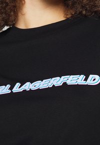 Camiseta de algodón negra con un destacado logo estilizado de "Karl Lagerfeld" en azul y rosa en la parte delantera, mangas cortas y cuello redondo.