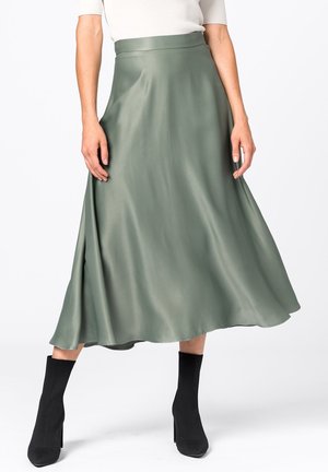 Kvinde iført en flydende midi-nederdel i olivengrøn, kombineret med sorte ankelstøvler med hæl og en hvid top.