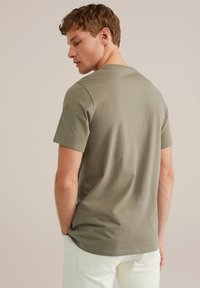 Olivno zelena bombažna majica s kratkimi rokavi, krožnim izrezom in sproščenim krojem, v kombinaciji z svetlimi hlačami.