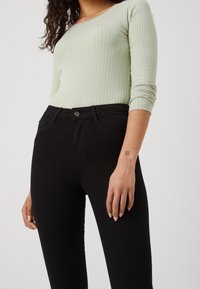 Woman wearing a light green ribbed long-sleeve top and high-waisted black skinny jeans, hand resting by her side.