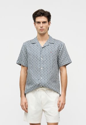 Junger Mann trägt ein kurzärmeliges, marineblau-weiß geometrisch gemustertes Hemd mit Kragen und weiße Shorts mit Kordelzug, steht vor einfachem Hintergrund.