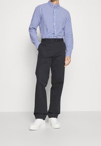 Sininen ja valkoinen ruudullinen nappipaita pitkähihaisilla hihoilla, yhdistettynä tummaan housuun ja valkoisiin lenkkarikenkiin, seisoo yksinkertaista taustaa vasten.