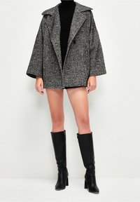 Grau und schwarzer Hahnentritt Oversized-Mantel mit weiten Ärmeln, großem Kragen und zwei Knöpfen, kombiniert mit kniehohen schwarzen Stiefeln mit Absatz.