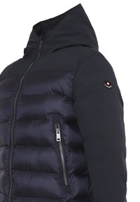 Giubbotto imbottito blu navy con un ampio cappuccio, dotato di una tasca laterale con zip e un logo rotondo sulla spalla sinistra. Texture liscia.