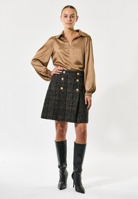 Blusa de satén marrón con cuello, combinada con una falda de cuadros negra con botones dorados y botas de tacón negras hasta la rodilla.