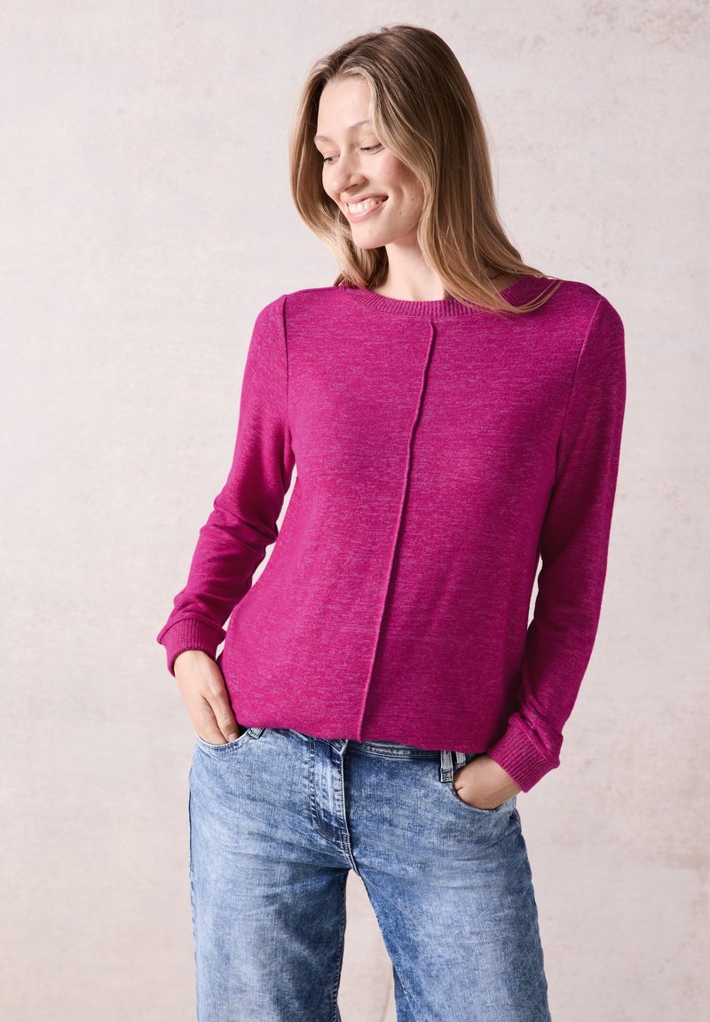 Pull en tricot fuchsia à manches longues, avec un détail de couture sur le devant. Associé à un jean en denim bleu clair et décontracté. Texture lisse.