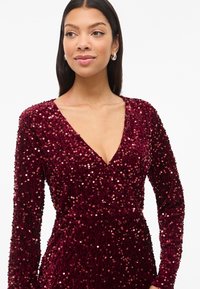 Bordová šatka zdobená flitrami s hlbokým výstrihom do V, dlhými rukávmi a textúrovaným povrchom; obsahuje lesklé flitre po celom povrchu.