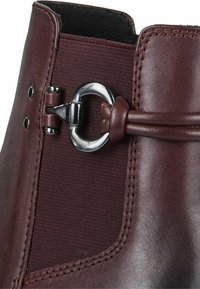 Botín de cuero marrón con panel de tela texturizada, herrajes metálicos y una hebilla decorativa con un detalle de cordones.