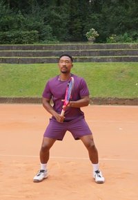 Joueur de tennis masculin en tenue violette, tenant une raquette de tennis avec un design rose et noir, sur un court en terre battue entouré de verdure.