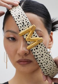 Ceinture en cuir beige avec des taches noires, dotée d'une boucle en métal doré torsadée au design en zigzag. Texture lisse avec une finition polie.