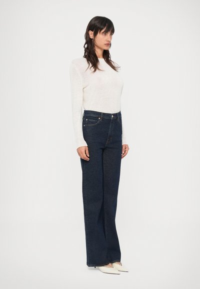 Σκούρα μπλε τζιν με ψηλή μέση και ίσια γραμμή, σε συνδυασμό με λευκό πλεκτό τοπ με μακριά μανίκια. Η υφή του υφάσματος φαίνεται απαλή και λεία.