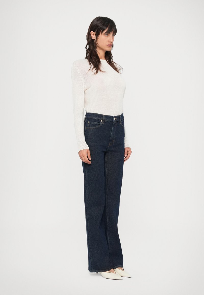 Dunkelblaue Jeans mit hoher Taille und geradem Bein, kombiniert mit einem weißen, langärmligen Stricktop. Die Stoffstruktur wirkt weich und glatt.