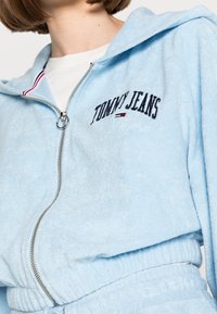Light blue zip-up hoodie in soft fabric, featuring embroidered "TOMMY JEANS" logo with red and white accent stitching on hood.