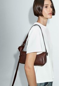 Borsa a tracolla in pelle marrone con una texture liscia, forma rettangolare e una tracolla corta, indossata sopra una camicia bianca. Presenta una chiusura con zip e dettagli in metallo.