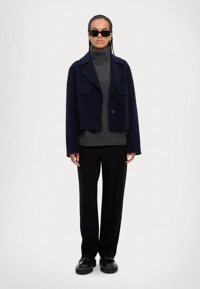 Marineblå crop-jakke med krave, to frontlommer, lagdelt over en mørkegrå ribbet turtleneck og sorte vide bukser.