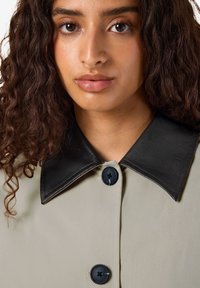 Vrouw met krullend bruin haar, gekleed in een beige jas met een grote zwart leren kraag en zwarte knopen, kijkt recht vooruit.