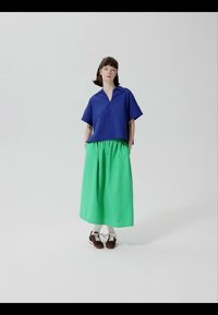 Kobieta stojąca, ubrana w niebieską koszulę z krótkim rękawem, zieloną długą spódnicę, białe skarpetki i brązowe buty sznurowane, na tle gładkiego jasnego tła.