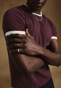 Bordo bombažna majica s kremnim robom, nošena s srebrnimi prstani na temni koži. Model prekrščene roke, ki poudarjajo teksturirano tkanino in nakit.