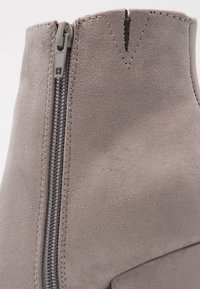 Gros plan sur une bottine en daim taupe avec une fermeture éclair verticale et des coutures détaillées près du bord supérieur.