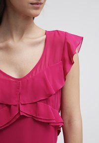 Blusa fucsia con volantes en capas, un escote en V profundo y mangas cortas de vuelo, hecha de un material ligero y fluido.