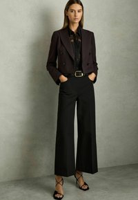 Zwarte dubbele-breasted cropped blazer over een zwart shirt, gecombineerd met wijde zwarte broek en zwarte hakken met bandjes. Over het geheel een gladde textuur.