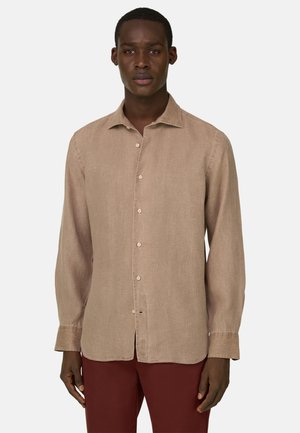 Mies, joka pukeutunut beigeen pitkähihainen nappipaitaan ja tummanpunaisiin housuihin, seisoo yksinkertaista vaaleaa taustaa vasten.