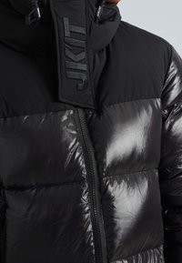 Svart puffjacka med glansig yta, vadderade sektioner, hög krage och logotypdetalj på dragkedjans flik.