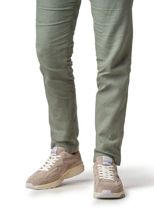Beige Wildleder-Sneaker mit weißen Schnürsenkeln und strukturiertem Design, kombiniert mit hellgrünen, schmal geschnittenen Hosen. Sichtbares Fersen- und Logodetail.
