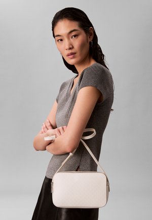 Žena sa ravnom tamnom kosom nosi sivu pletenu majicu kratkih rukava i drži malu bijelu torbicu preko tijela ispred jednostavne sive pozadine.
