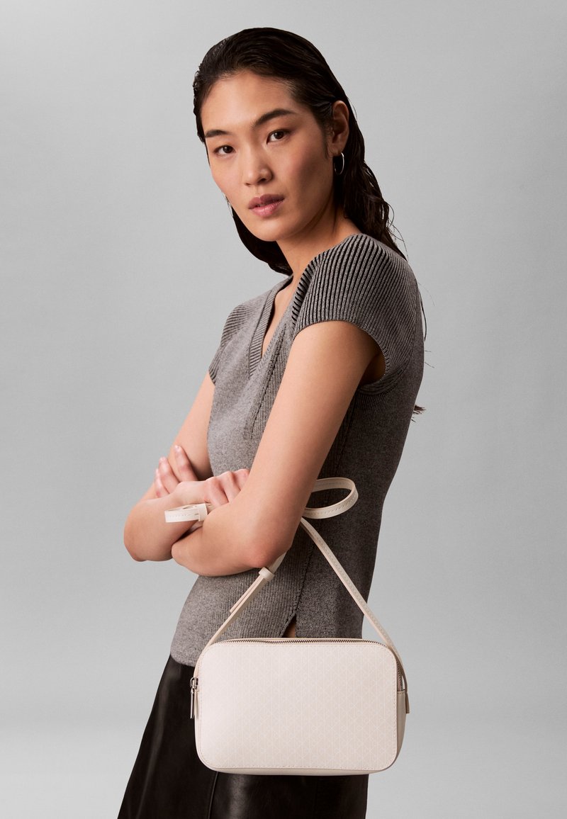 Sirgete tumedate juustega naine kannab halli lühikeste varrukatega kootud toppi ning hoiab käes väikest valget ristiirukkuga kotti tavalise halli tausta ees.