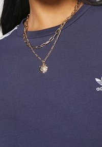 Tmavě modré tričko s krátkým rukávem a žebrovaným výstřihem, zdobené vrstvenými řetězy ve zlatém tónu a náhrdelníkem se slunečním pendantem.