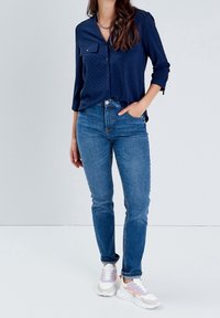 Femme portant une chemise boutonnée bleu marine, un jean bleu et des baskets blanches avec des accents rose et beige, se tenant avec une main dans la poche.