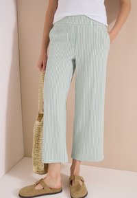 Woman wearing light green and white striped wide-leg pants, beige slip-on shoes, and holding a woven tote bag by her side.