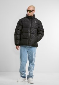 Sort pufferjakke med høj krave, udstyret med subtile mønstre, parret med lyseblå slidte jeans og hvide sneakers.