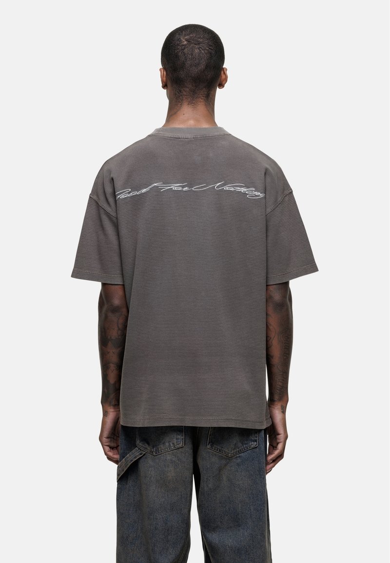 Grijze oversized t-shirt met korte mouwen en een geribbelde textuur. Heeft witte cursieve tekst op de achterkant. Gecombineerd met denimbroeken.