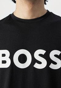 Henkilö, joka käyttää mustaa pyöreäkaulaista paitaa, jossa on suuret valkoiset kirjaimet, jotka kirjoittavat "BOSS" rinnan yli, lähikuva ylävartalosta ja kaulasta.