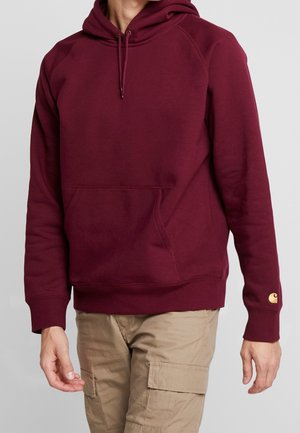 Person trägt einen burgunderfarbenen Kapuzenpullover mit Fronttasche und beigen Cargohosen, steht vor einem schlichten grauen Hintergrund.