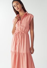 Sukienka zapinana na guziki w miękkim koralowym materiale, z kołnierzykiem, krótkimi rękawami, elastycznym paskiem w talii oraz warstwową, zwiewną spódnicą.