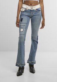 Jean en denim bleu clair avec une coupe évasée, accents déchirés, détails en patchwork et ourlet effiloché. Taille blanche avec fermeture à bouton.