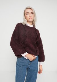 Burgundowy, fakturowany sweter z falbaniastymi akcentami, okrągłym dekoltem i opadającymi ramionami, noszony na różowej koszuli z kołnierzykiem, w zestawieniu z niebieskimi dżinsami.