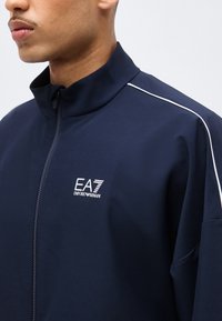 Άτομο που φοράει μπλε σκούρο μπουφάν με φερμουάρ, με λευκό λογότυπο EA7 Emporio Armani στο στήθος και λευκή διακοσμητική ραφή στον ώμο.