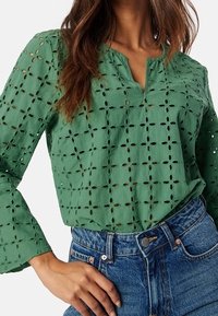 Blouse verte à manches longues avec broderie ajourée et décolleté en V, associée à un jean bleu taille haute. Tissu texturé avec une coupe ample.