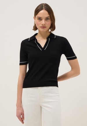 Vrouw met een zwart poloshirt met korte mouwen en witte bies op de kraag en mouwen, gecombineerd met witte broek met hoge taille.