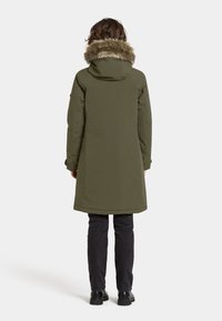 Olivengrøn parka med en pelsforet hætte, der har en mellemlang silhuet og et strømlinet design. Kombineret med slanke sorte bukser og sorte sko.
