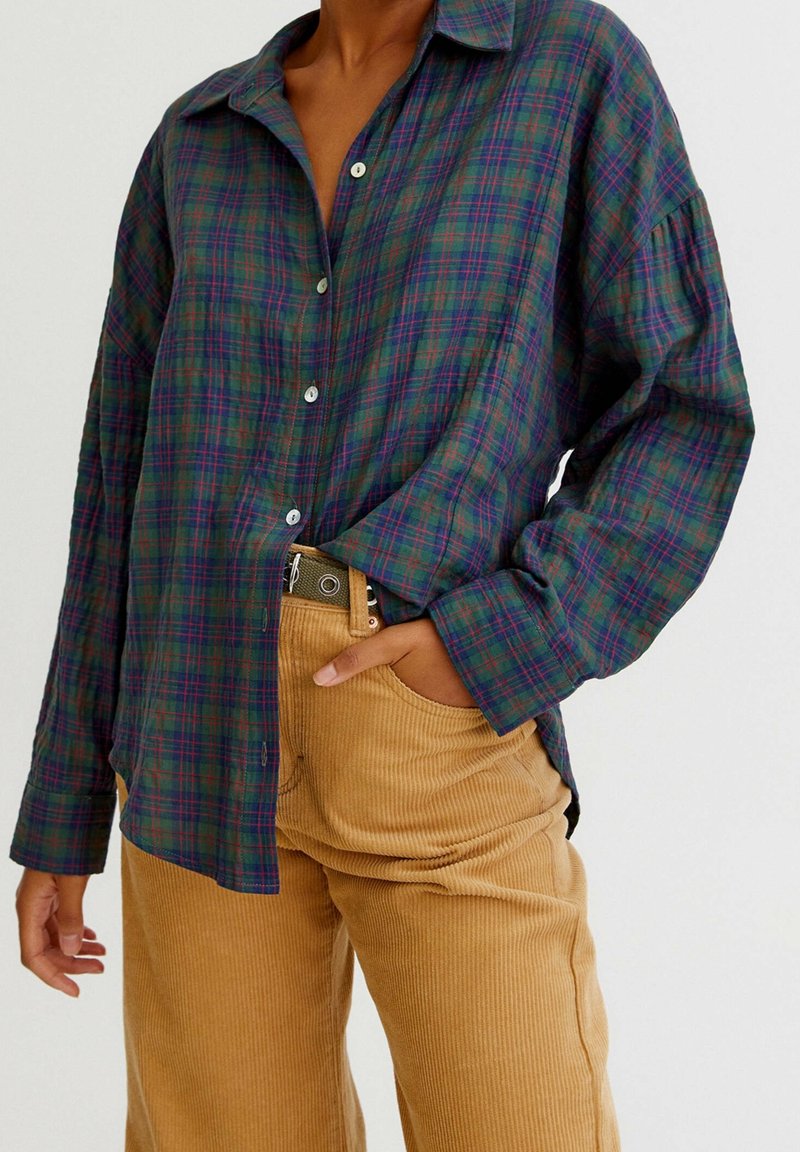 Chemise à manches longues en flanelle à carreaux vert foncé et bleu marine, dotée d'un col pointu, d'une fermeture à boutons et d'une coupe décontractée. Associée à un pantalon en velours côtelé beige.