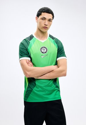 Jonge man met kort zwart haar, gekleed in een groen en donkergroen Nigeria Football Federation-shirt, met gekruiste armen op een witte achtergrond.