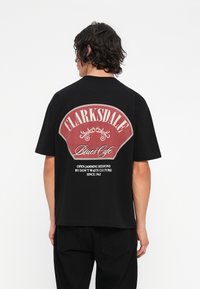 Camiseta de algodón negra con un gran gráfico en rojo y blanco en la parte posterior que dice "Clarksdale Blues Cafe" con elementos decorativos.