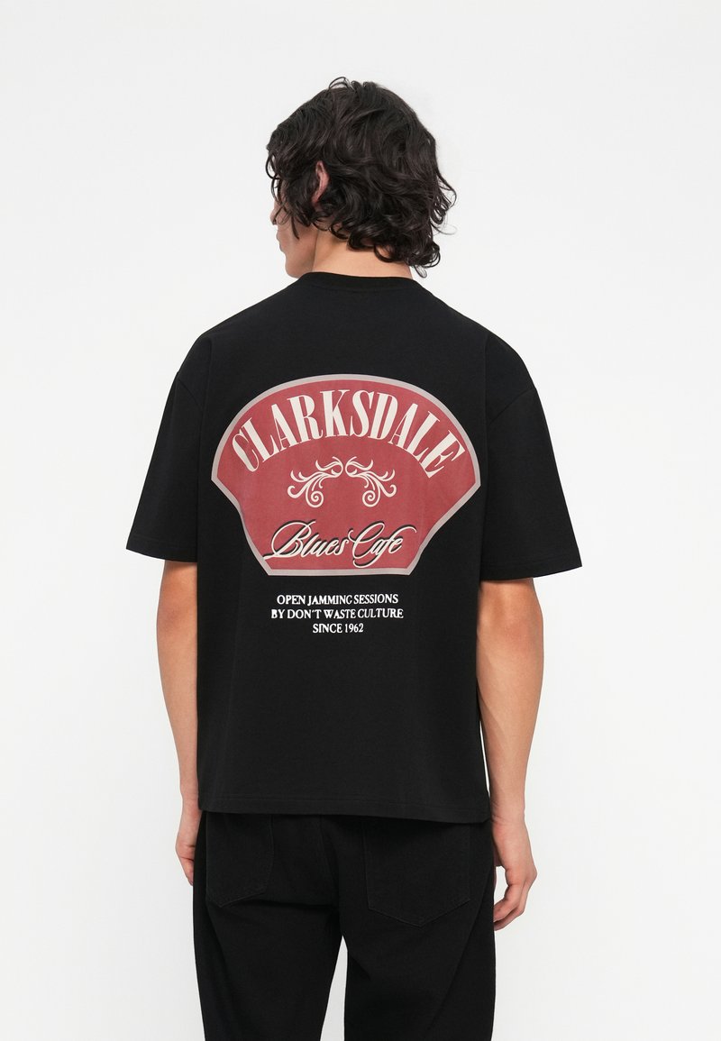 Camiseta de algodón negra con un gran gráfico en rojo y blanco en la parte posterior que dice "Clarksdale Blues Cafe" con elementos decorativos.